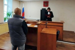 Мужчина стоит перед судьёй, читающей документ у стола в зале суда с российским флагом и гербом.